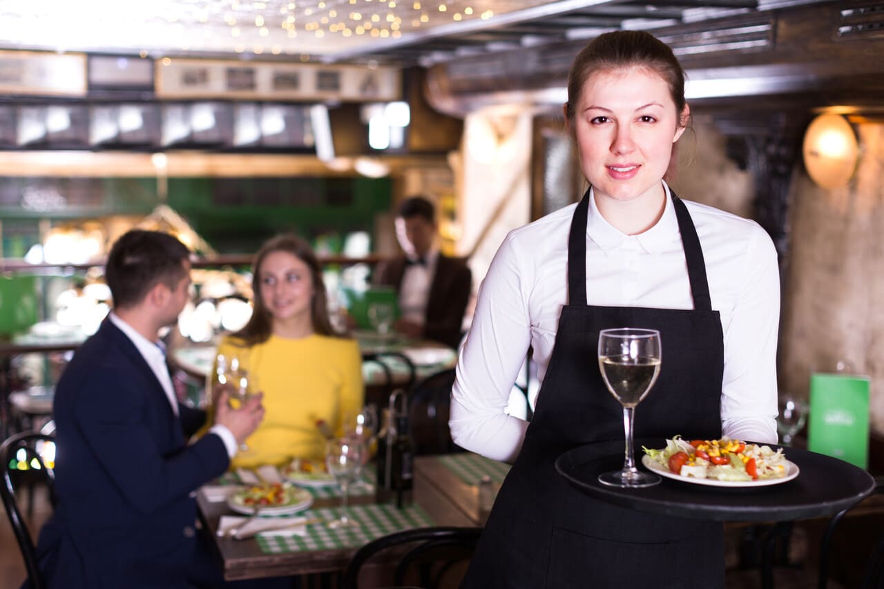 Woman waiter is holding tray with wine and salad for clients in restaurant.