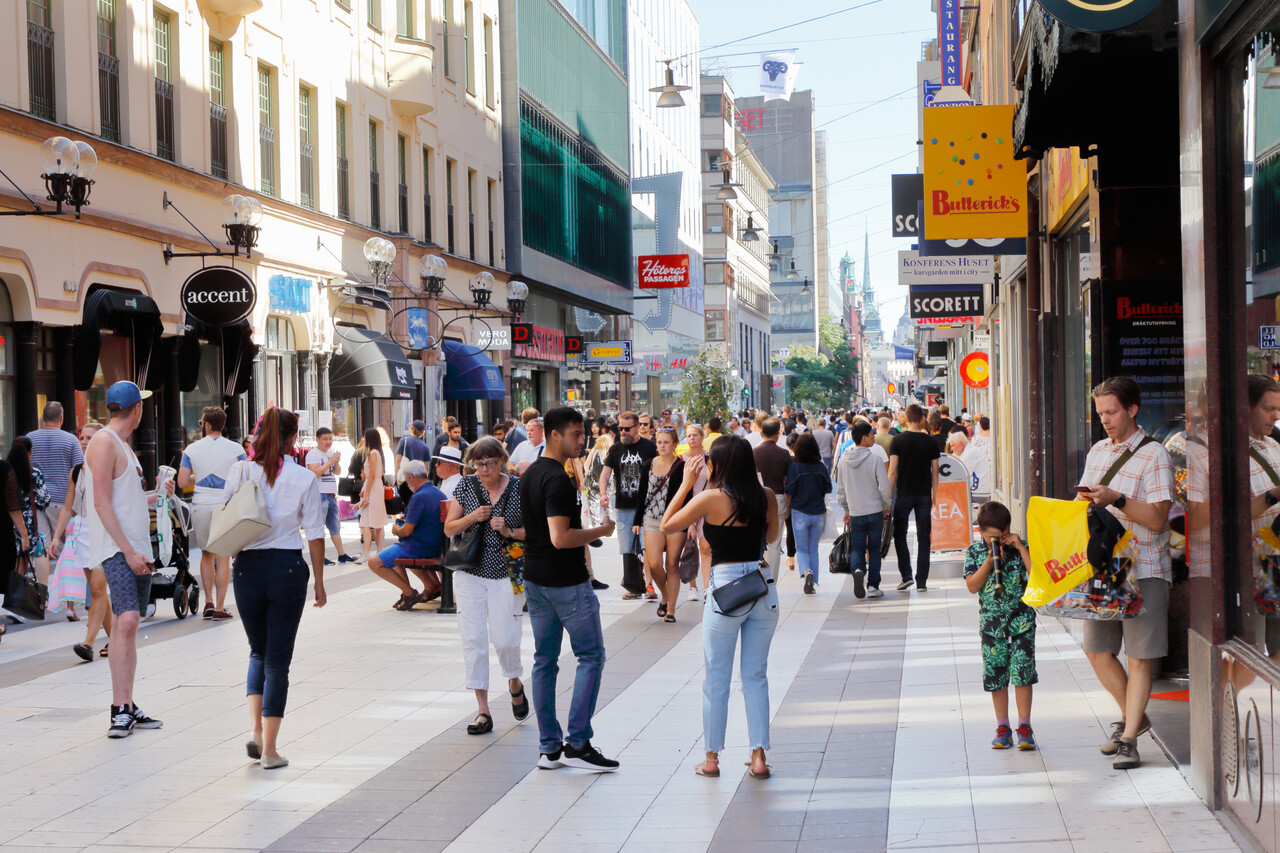 Crowded tourist street scene where one person distracts a traveler while another reaches toward an open backpack or pocket.