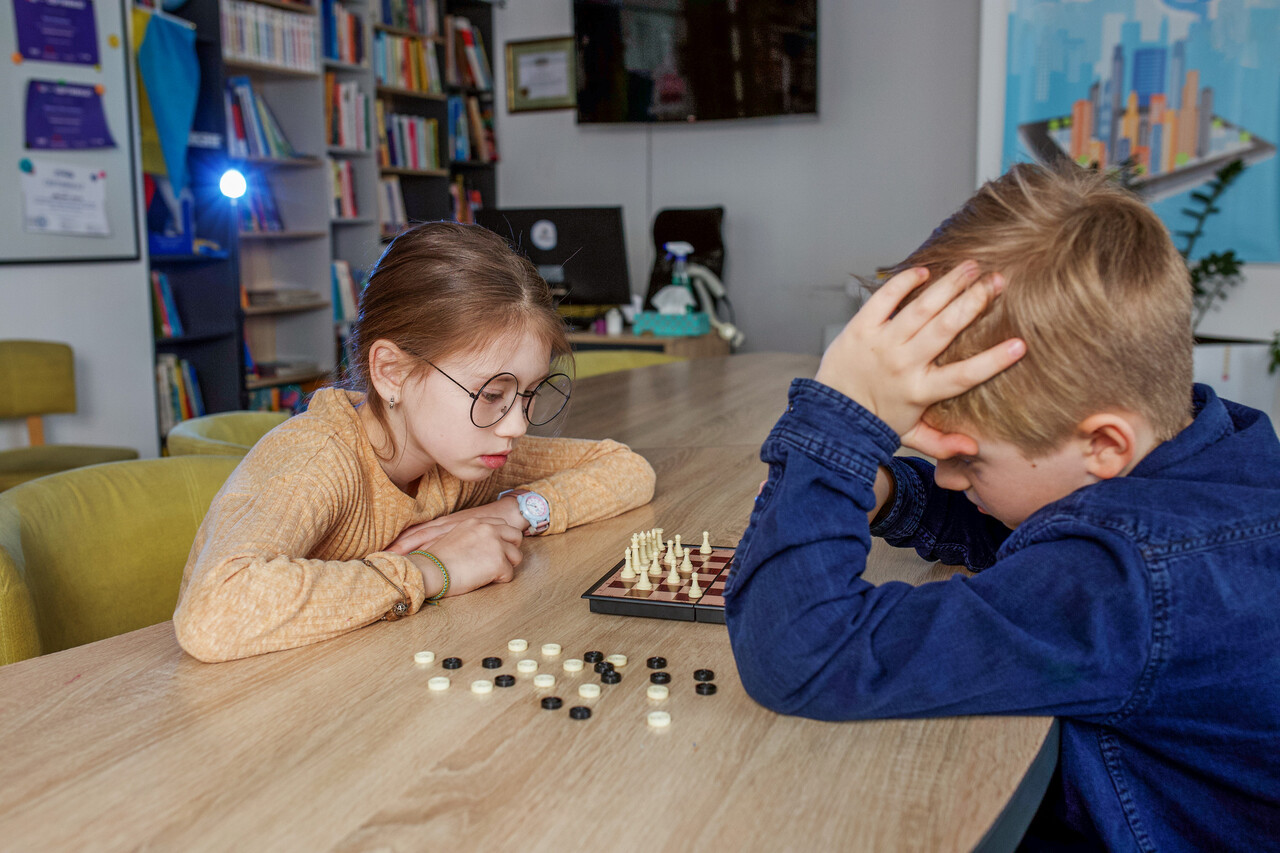 A wooden chessboard mid-game with two students concentrating, photographed with soft classroom lighting.