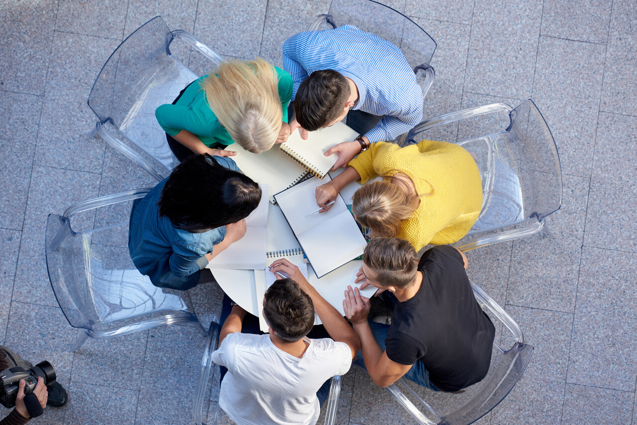Students seated in a circle with newspapers and notepads, one speaking while others listen.