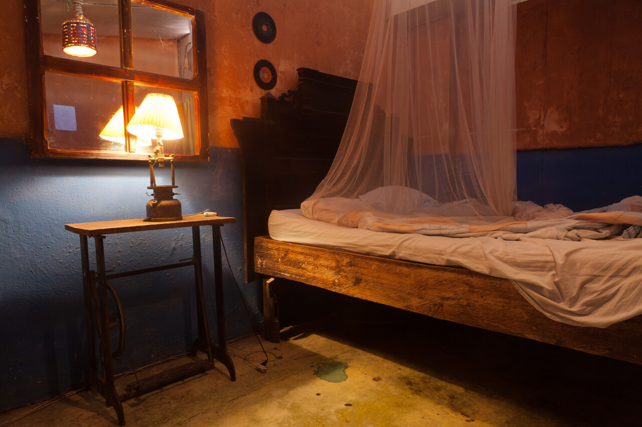 A dimly lit old inn room with a wooden door slightly ajar and a simple bed turned away from it.