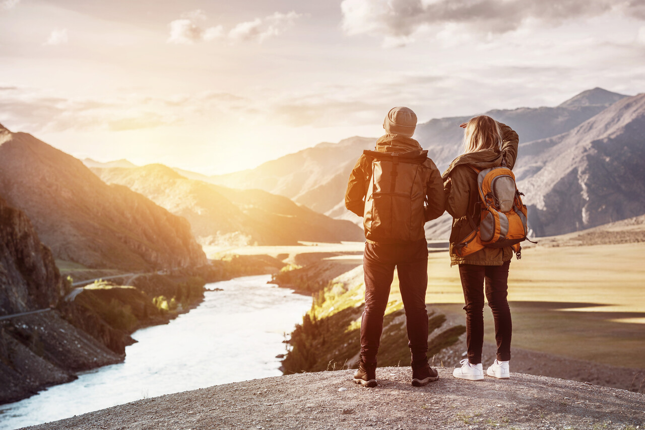 A wide, warm-toned photo of a couple with backpacks looking out over a sweeping landscape (mountains, coast, or countryside).