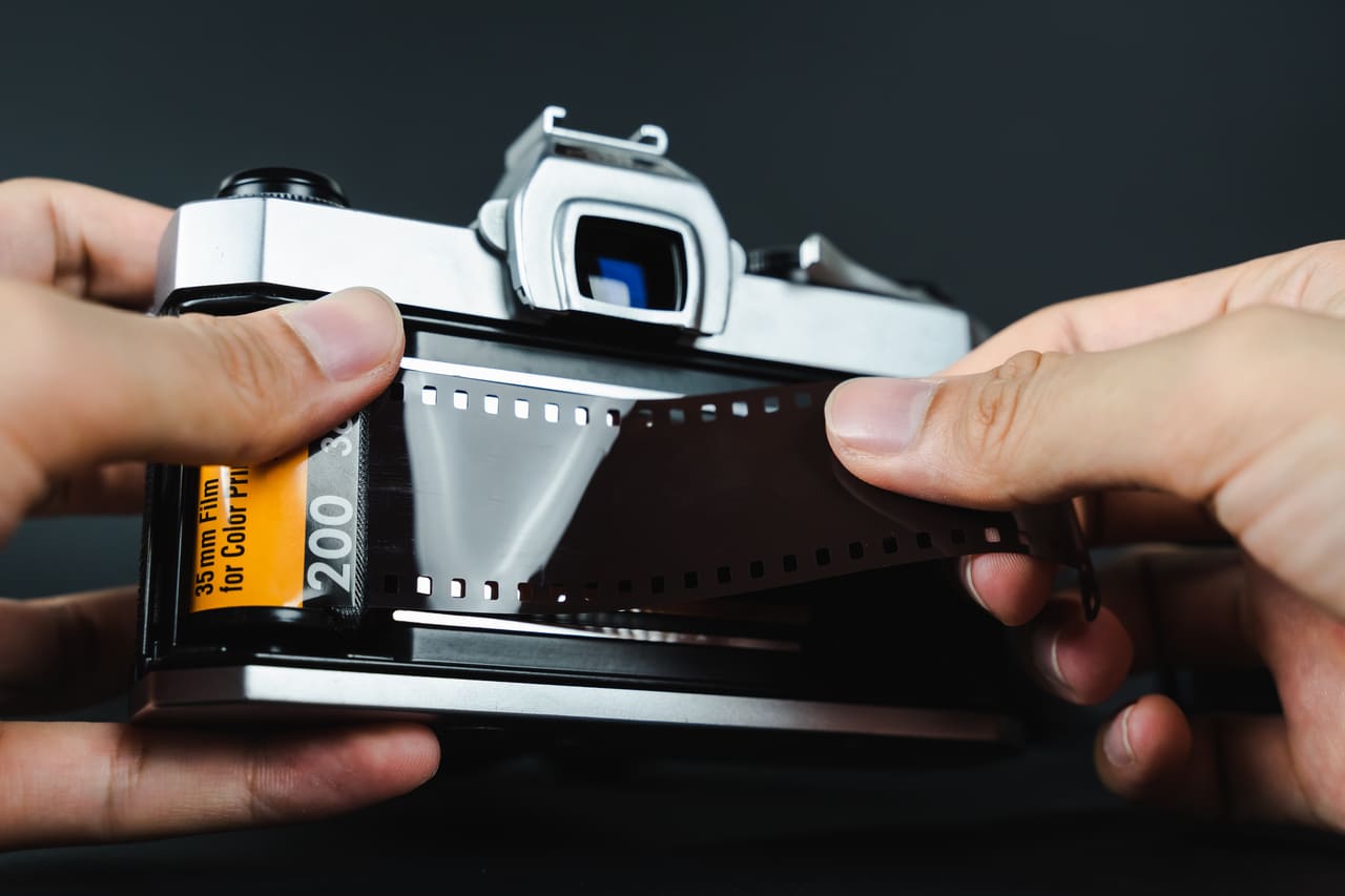 A student holding a 35mm film camera or working in a dim darkroom with trays and drying prints.