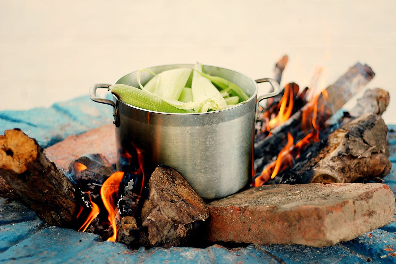 A clay pot over an open flame with vegetables roasting inside, steam rising.